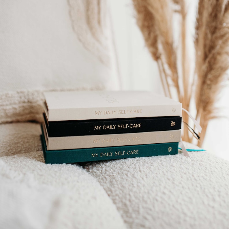 May include: A stack of four journals with the text "My Daily Self-Care" on the front cover. The journals are in different colours: cream, black, cream, and teal.