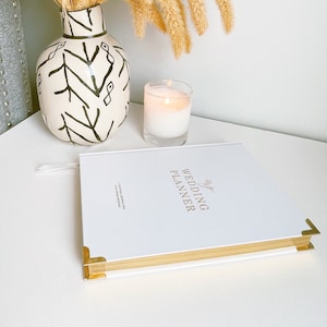 K&ouml;nnte beinhalten: Ein wei&szlig;er Hochzeitsplaner mit goldenen Akzenten und dem Text "My Wedding Planner" auf dem Cover. Der Planer liegt auf einer wei&szlig;en Oberfl&auml;che mit einer brennenden Kerze und einer Vase mit einem schwarz-wei&szlig;en Muster.