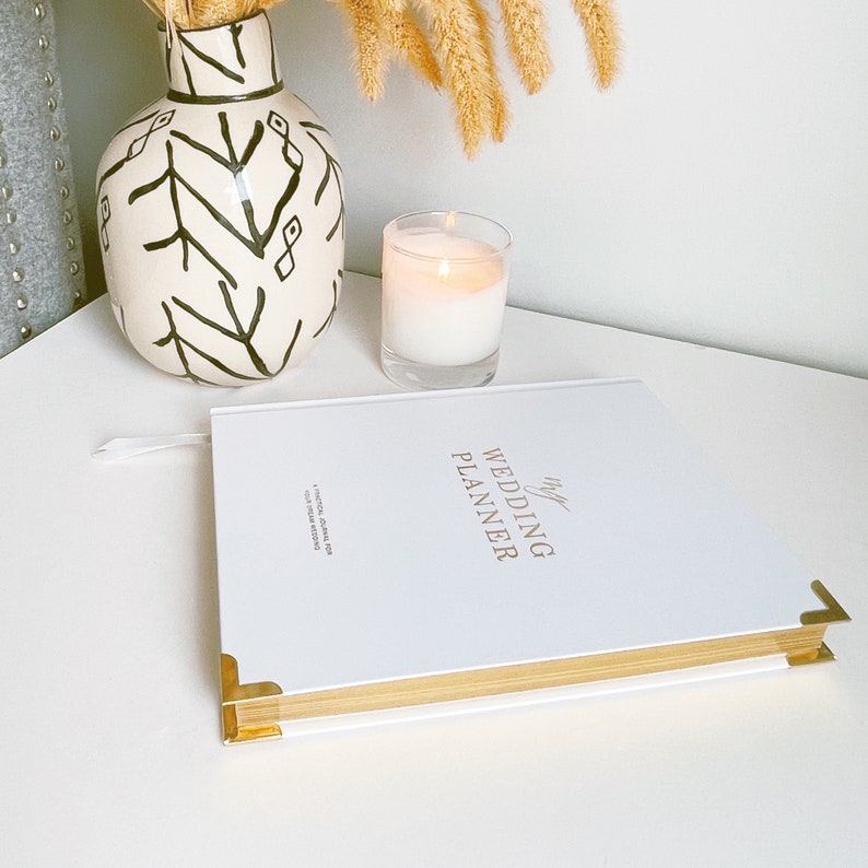 K&ouml;nnte beinhalten: Ein wei&szlig;er Hochzeitsplaner mit goldenen Akzenten und dem Text "My Wedding Planner" auf dem Cover. Der Planer liegt auf einer wei&szlig;en Oberfl&auml;che mit einer brennenden Kerze und einer Vase mit einem schwarz-wei&szlig;en Muster.