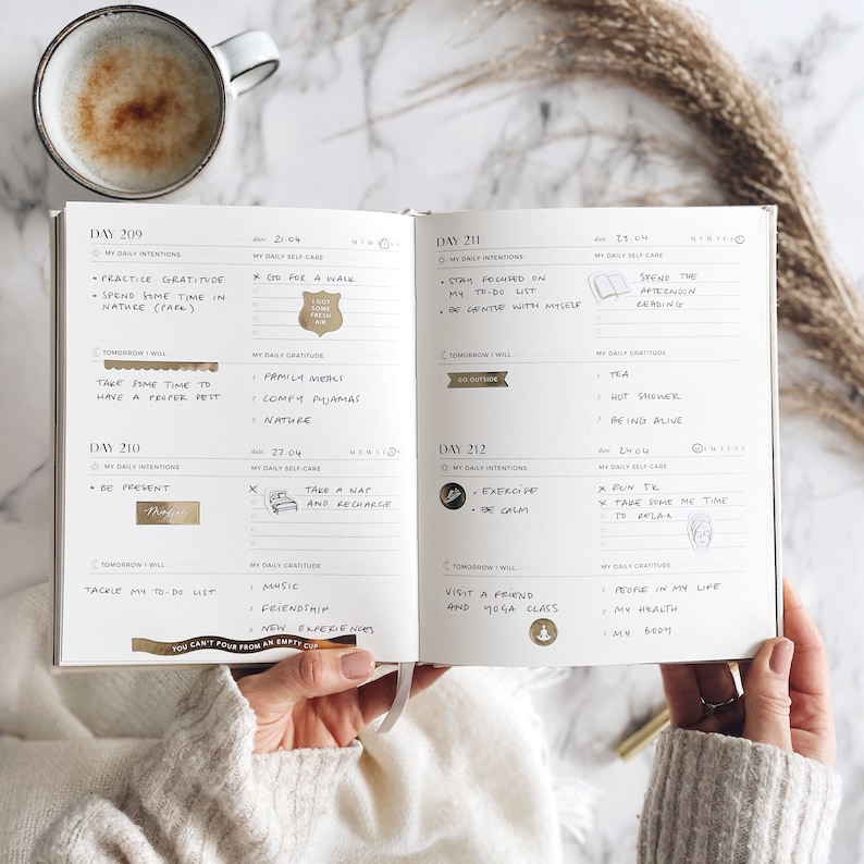 Op de afbeelding: Een open planner met dagelijkse intenties en doelen voor zelfzorg, geschreven in gouden letters. De planner heeft een witte achtergrond en is versierd met gouden stickers. De planner is open op de pagina's voor dag 209, 210, 211 en 212.