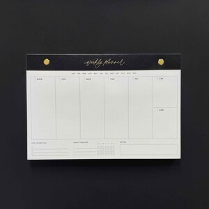 K&ouml;nnte beinhalten: Ein schwarz-wei&szlig;er Wochenplaner mit goldenen Akzenten. Der obere Teil des Planers ist schwarz mit dem goldfarbenen Schriftzug "weekly planner". Er hat Abschnitte f&uuml;r jeden Wochentag, Top-Priorit&auml;ten, einen Gewohnheitstracker und Notizen.