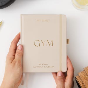 Puede incluir: Una libreta beige con un título dorado "MY DAILY GYM" y el texto "the ultimate WORKOUT LOGBOOK".