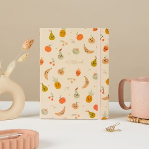 May include: A cream-coloured notebook with a fun fruit pattern. The cover features a variety of fruits, including pears, bananas, pineapples, strawberries, and cherries, all with cute faces. The notebook has a pink elastic closure and the word "notebook" is written in cursive on the front.