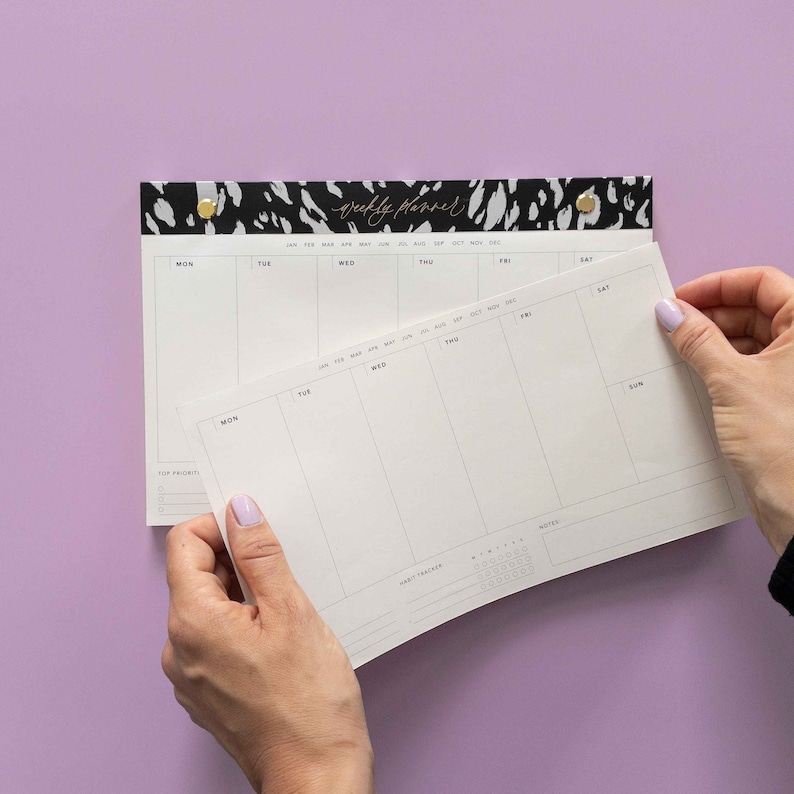 May include: A white weekly planner with a black and white animal print design on the top. The planner has sections for each day of the week, with space for notes and a habit tracker.