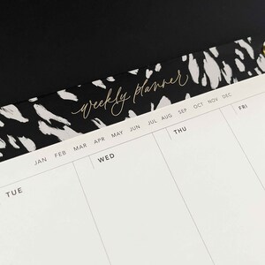 May include: A white weekly planner with black and white abstract animal print design on the top. The planner has a column for each day of the week, with months listed across the top.