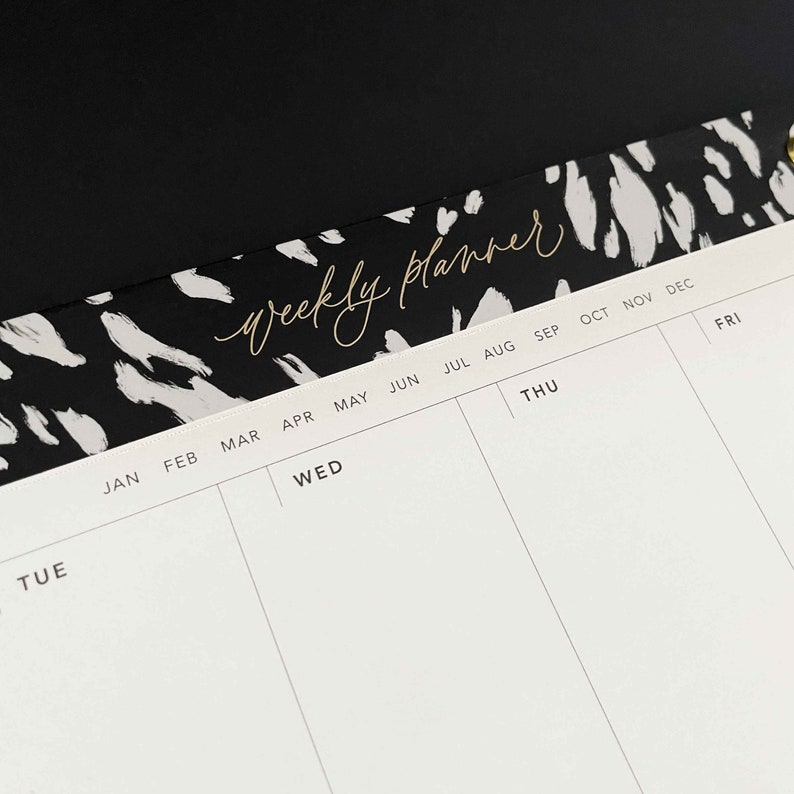 May include: A white weekly planner with black and white abstract animal print design on the top. The planner has a column for each day of the week, with months listed across the top.