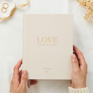 May include: A cream-coloured wedding planner with the word "LOVE" in gold lettering. The text "THE WEDDING PLANNER" is below the main title. The phrase "for all couples" is printed at the bottom. The planner is held by two hands.