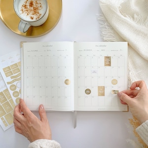 K&ouml;nnte beinhalten: Offener Planer mit goldenen Aufklebern auf den Seiten. Der Planer hat einen wei&szlig;en Hintergrund und die Monate September und November sind sichtbar. Die Aufkleber haben Goldfolie und sagen "Good Vibes" und "Today Found Green".