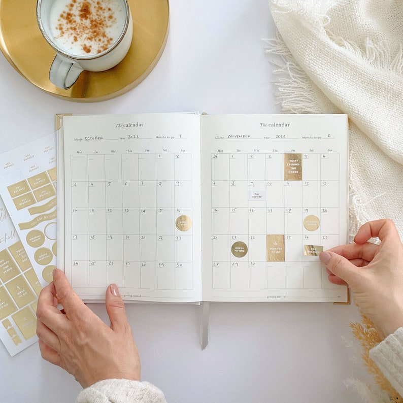 K&ouml;nnte beinhalten: Offener Planer mit goldenen Aufklebern auf den Seiten. Der Planer hat einen wei&szlig;en Hintergrund und die Monate September und November sind sichtbar. Die Aufkleber haben Goldfolie und sagen "Good Vibes" und "Today Found Green".