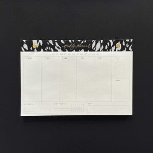May include: A white weekly planner with a black and white animal print design at the top. The planner has sections for each day of the week, a habit tracker, and a notes section. The text "weekly planner" is written in gold cursive lettering.