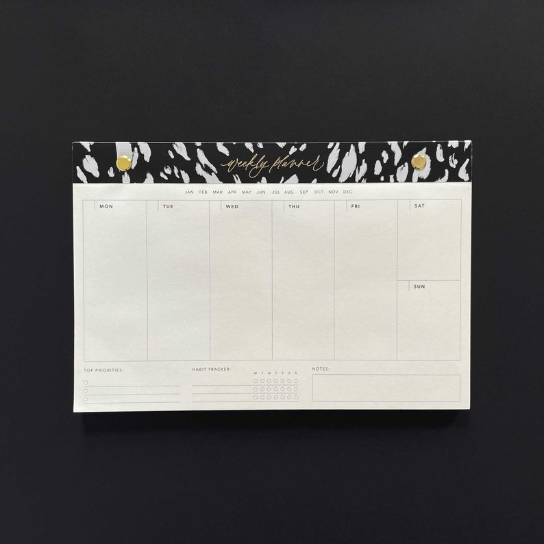 May include: A white weekly planner with a black and white animal print design at the top. The planner has sections for each day of the week, a habit tracker, and a notes section. The text "weekly planner" is written in gold cursive lettering.