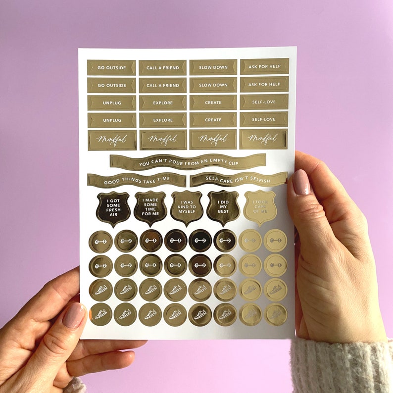 May include: Gold foil stickers with motivational phrases and illustrations. The stickers include phrases like "Go Outside", "Call a Friend", "Slow Down", "Ask for Help", "Unplug", "Explore", "Create", "Self-Love", "You Can't Pour From an Empty Cup", "Good Things Take Time", "Self-Care Isn't Selfish", "I Got Some Fresh Air", "I Made Some Time For Me", "I Was Kind to Myself", "I Did My Best", and "I Took Care of Me". The stickers also include illustrations of a dumbbell, a shoe, and a shield.