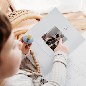 Pu&ograve; includere: Un bambino tiene in mano una catena per ciuccio blu e gialla e guarda un album fotografico. L'album &egrave; aperto a una pagina con una foto di una donna incinta e il testo "settimana 23".