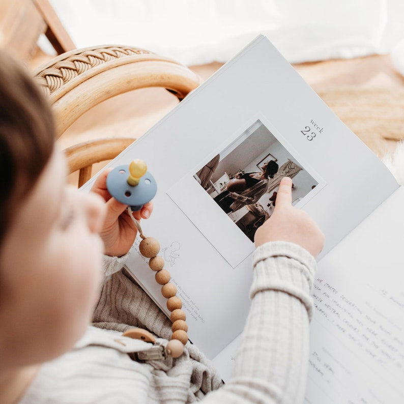 Pu&ograve; includere: Un bambino tiene in mano una catena per ciuccio blu e gialla e guarda un album fotografico. L'album &egrave; aperto a una pagina con una foto di una donna incinta e il testo "settimana 23".