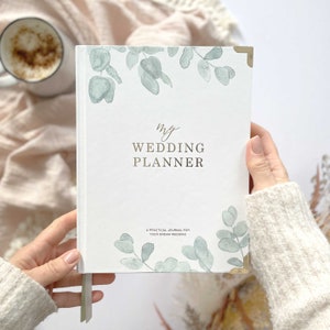 May include: A white wedding planner with a gold foil title "my WEDDING PLANNER" and a green watercolour floral design. The text "A PRACTICAL JOURNAL FOR YOUR DREAM WEDDING" is printed below the title.