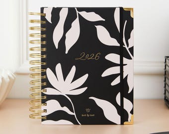 May include: A black and white floral 2026 planner with gold spiral binding and corner protectors. The cover features a repeating pattern of white leaves and flowers. The words "2026" and "DAY BY DAY" are printed on the cover.