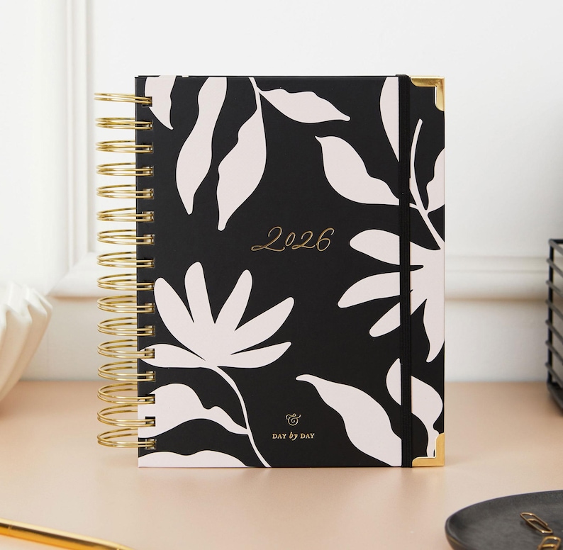 May include: A black and white floral 2026 planner with gold spiral binding and corner protectors. The cover features a repeating pattern of white leaves and flowers. The words "2026" and "DAY BY DAY" are printed on the cover.
