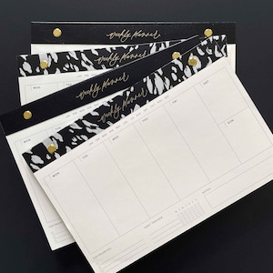 K&ouml;nnte beinhalten: Mehrere Wochenplaner mit einem schwarz-wei&szlig;en Tiermuster. Auf der Oberseite jedes Planers steht in goldener Schrift "weekly planner". Die Planer sind mit goldfarbenen Nieten befestigt und liegen auf einer schwarzen Oberfl&auml;che.