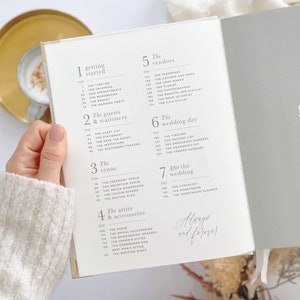K&ouml;nnte beinhalten: Ein Hochzeitsplaner mit wei&szlig;em Einband und goldenen Akzenten. Das Buch ist auf einer Seite mit dem Titel "Getting Started" ge&ouml;ffnet und listet verschiedene Aufgaben f&uuml;r die Hochzeitsplanung auf, wie z. B. den Zeitplan, den Kalender, Termine und das Budget.