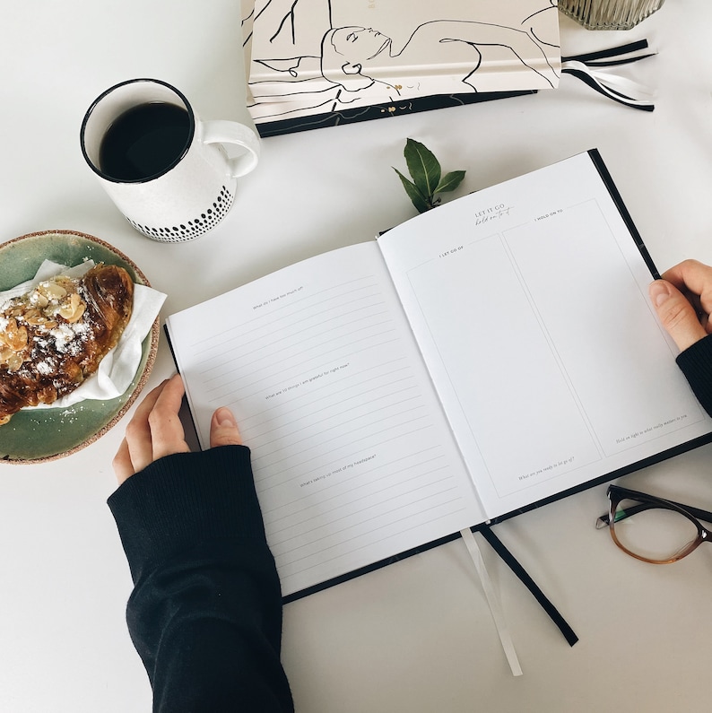 May include: An open black journal with the text "LET IT GO hold on to it" is held open. A cup of coffee, a croissant, and a decorative book are also on the white surface. The journal has a black ribbon bookmark.
