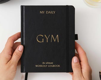 Puede incluir: Diario encuadernado en cuero negro con letras doradas que dicen "MY DAILY GYM the ultimate WORKOUT LOGBOOK".