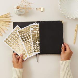 May include: A black journal with gold corner accents and a black ribbon bookmark. Gold stickers with various labels and shapes are held next to the journal. A gold pen, binder clip, and decorative items are also on the cream-colored surface.