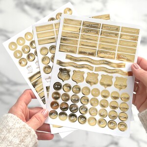 May include: A collection of gold foil stickers with various motivational phrases and icons. The stickers include text like "Digital Break," "Go Outside," and "Self Love," along with images of dumbbells and sneakers. The stickers are held in a person's hands.