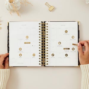 May include: An open black and gold spiral-bound planner with the month of August displayed. The planner is open to a calendar view with gold foil stickers and handwritten notes. A gold paper clip rests on the surface.