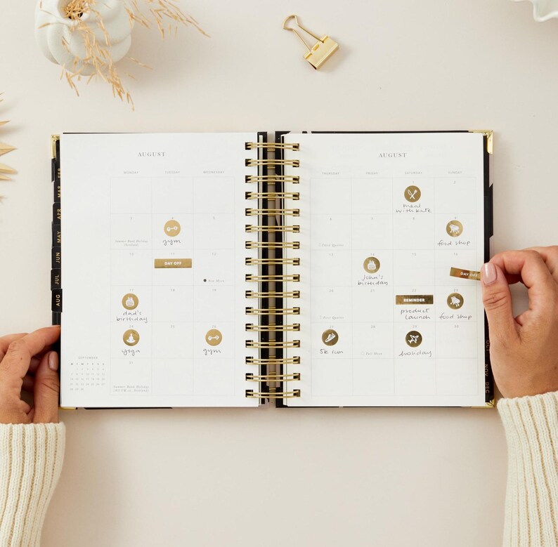 May include: An open black and gold spiral-bound planner with the month of August displayed. The planner is open to a calendar view with gold foil stickers and handwritten notes. A gold paper clip rests on the surface.
