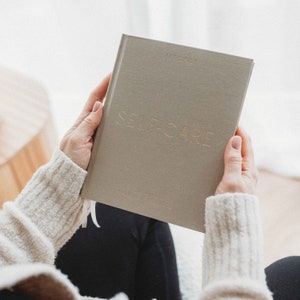 May include: A grey self-care journal with gold lettering. The journal is being held by a person wearing a white sweater.