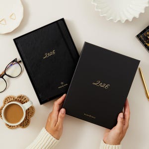 Peut inclure: Deux agendas noirs de 2026 avec des lettres dor&eacute;es et une bo&icirc;te assortie. Un agenda est ouvert, r&eacute;v&eacute;lant les pages int&eacute;rieures. L'autre agenda est ferm&eacute;. Les mots "Week by Week" et "Blush & Gold" sont imprim&eacute;s sur les agendas.