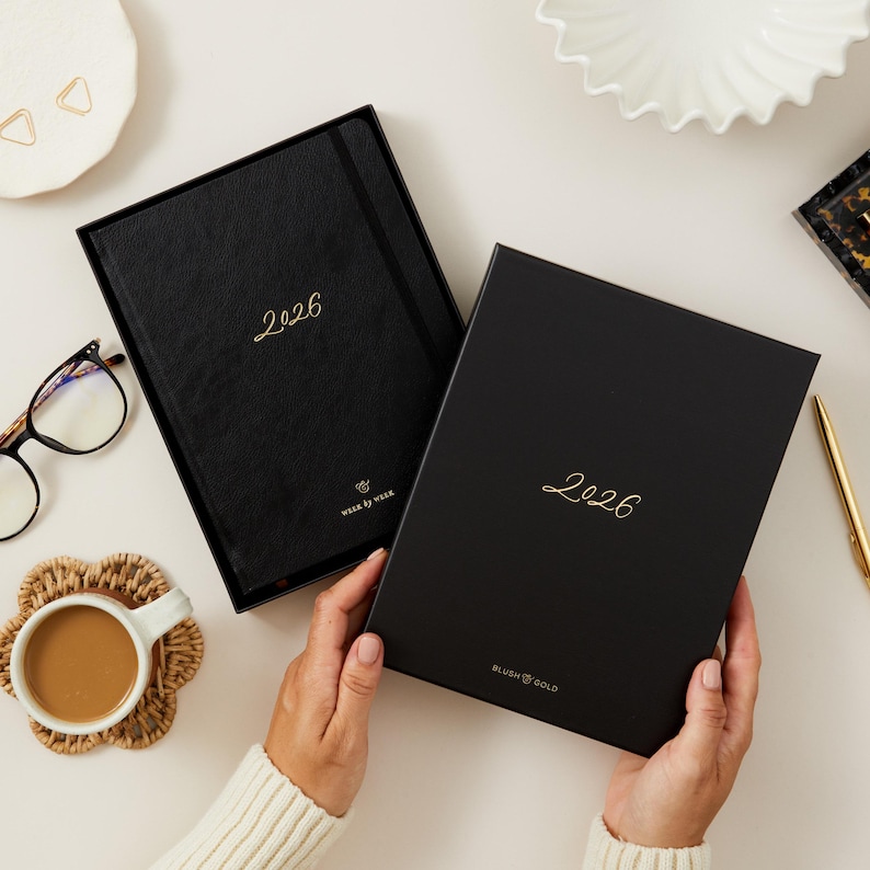 Peut inclure: Deux agendas noirs de 2026 avec des lettres dor&eacute;es et une bo&icirc;te assortie. Un agenda est ouvert, r&eacute;v&eacute;lant les pages int&eacute;rieures. L'autre agenda est ferm&eacute;. Les mots "Week by Week" et "Blush & Gold" sont imprim&eacute;s sur les agendas.
