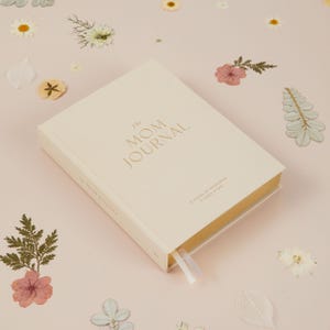 May include: A cream-colored journal titled "The MOM JOURNAL" with gold lettering. The journal has a cream-colored ribbon bookmark and the text "5 YEARS OF MEMORIES & LINES A DAY" is printed on the cover. The journal is on a pink surface with dried flowers.
