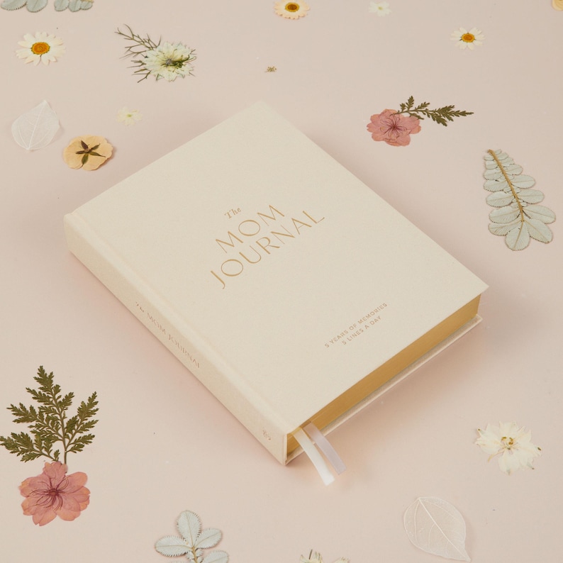 May include: A cream-colored journal titled "The MOM JOURNAL" with gold lettering. The journal has a cream-colored ribbon bookmark and the text "5 YEARS OF MEMORIES & LINES A DAY" is printed on the cover. The journal is on a pink surface with dried flowers.