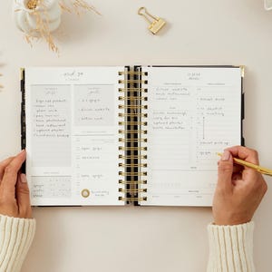 May include: An open, spiral-bound planner with gold accents, featuring handwritten notes and a gold pen. The planner is open to a weekly spread with sections for tasks and goals. The text "29" and "IV JULY" are visible.