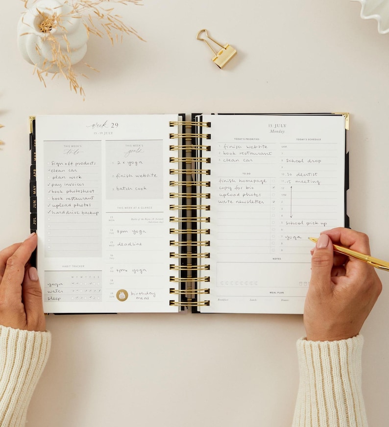 May include: An open, spiral-bound planner with gold accents, featuring handwritten notes and a gold pen. The planner is open to a weekly spread with sections for tasks and goals. The text "29" and "IV JULY" are visible.