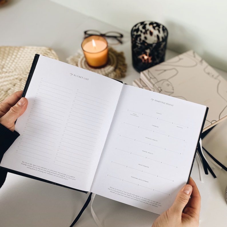 May include: Open black journal with white pages, featuring a "Bucket List" and "Starting Point" section. The journal is held open, revealing lined pages for notes and goal setting. A lit candle and decorative items are in the background.