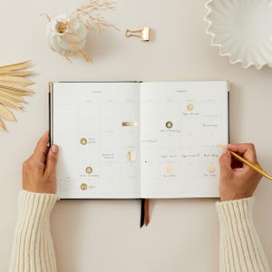 Peut inclure: Un agenda ouvert avec un calendrier de mars, des d&eacute;tails dor&eacute;s et des notes manuscrites. Une personne &eacute;crit dans l'agenda avec un stylo dor&eacute;. L'agenda est pos&eacute; sur une surface claire, avec des &eacute;l&eacute;ments d&eacute;coratifs en arri&egrave;re-plan.