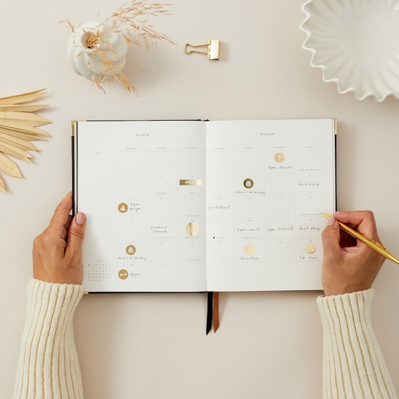 Peut inclure: Un agenda ouvert avec un calendrier de mars, des d&eacute;tails dor&eacute;s et des notes manuscrites. Une personne &eacute;crit dans l'agenda avec un stylo dor&eacute;. L'agenda est pos&eacute; sur une surface claire, avec des &eacute;l&eacute;ments d&eacute;coratifs en arri&egrave;re-plan.