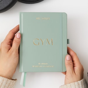 May include: A sage green journal with a gold embossed title "MY DAILY GYM" and the text "the ultimate workout log for your" in gold lettering.