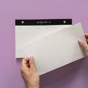 K&ouml;nnte beinhalten: Ein schwarzer und goldener Wochenplaner mit einem wei&szlig;en Papiereinsatz. Der Planer tr&auml;gt den Schriftzug "weekly planner" in goldener Schrift. Der Einsatz wird von einer Person mit hellviolettem Nagellack gehalten. Der Hintergrund ist einfarbig lila.