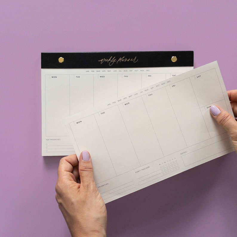 K&ouml;nnte beinhalten: Ein schwarzer und goldener Wochenplaner mit einem wei&szlig;en Papiereinsatz. Der Planer tr&auml;gt den Schriftzug "weekly planner" in goldener Schrift. Der Einsatz wird von einer Person mit hellviolettem Nagellack gehalten. Der Hintergrund ist einfarbig lila.