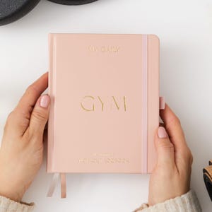May include: A pink daily gym workout logbook with a gold embossed "GYM" on the front cover.