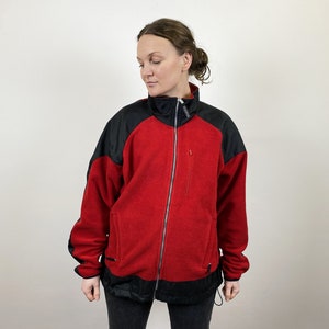 Könnte beinhalten: Eine rote Fleecejacke mit schwarzen Akzenten. Die Jacke hat einen Reißverschluss und zwei Taschen.