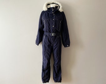 Könnte beinhalten: Ein dunkelblauer Ski-Overall mit einem weißen Kunstfellbesatz an der Kapuze. Der Overall hat einen Reißverschluss, einen Gürtel und weiße Nähte.
