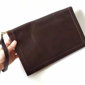 Puede incluir: Un clutch de cuero marrón con un cierre dorado y una correa. El clutch tiene una superficie lisa y texturizada.