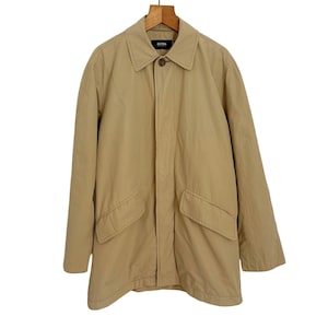 May include: A tan-colored coat with a collar and two front pockets hangs from a wooden hanger. The coat has a button closure and a simple, classic design. The coat is made of a lightweight material.