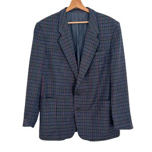 Puede incluir: Un blazer a cuadros azul, rojo y negro colgado de una percha de madera. El blazer tiene cierre de dos botones, dos bolsillos delanteros y solapa con muescas. El forro interior es de color azul liso.