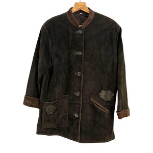 Könnte beinhalten: Dunkelbraune Wildlederjacke mit rundem Kragen und Knopfverschluss. Mit zwei Vordertaschen und blattförmigen Applikationen. Die Manschetten und der Kragen sind in einem dunkleren Braunton gehalten.
