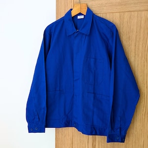 Könnte beinhalten: Eine leuchtend königsblaue Arbeitsjacke mit langen Ärmeln, klassischem Kragen, einer Brusttasche, zwei unteren Vordertaschen und geknöpften Manschetten. Die Jacke hat eine Knopfleiste und hängt an einem hölzernen Kleiderbügel.